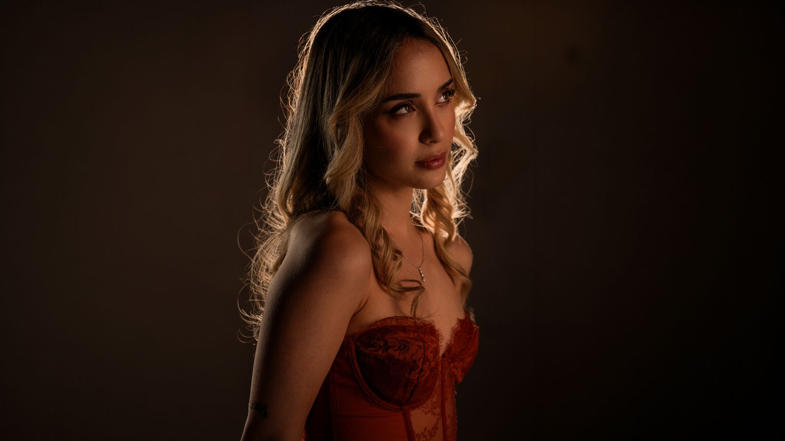 Raphaela in a red lace corset, backlit against a dark backdrop and looking off to the side, press photo, 2026.
